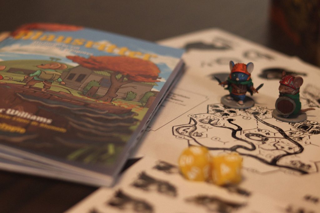 Libro de Mausritter junto a miniaturas de ratones aventureros, dados amarillos y un mapa dibujado en blanco y negro sobre la mesa.