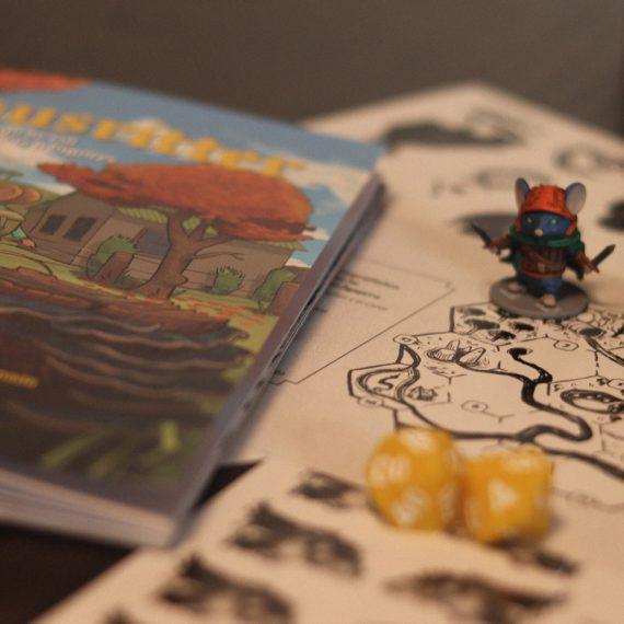 Libro de Mausritter junto a miniaturas de ratones aventureros, dados amarillos y un mapa dibujado en blanco y negro sobre la mesa.