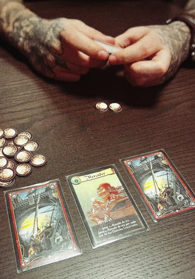 Manos jugando a Ciudadela, con cartas y monedas sobre la mesa durante una partida de juego de mesa
