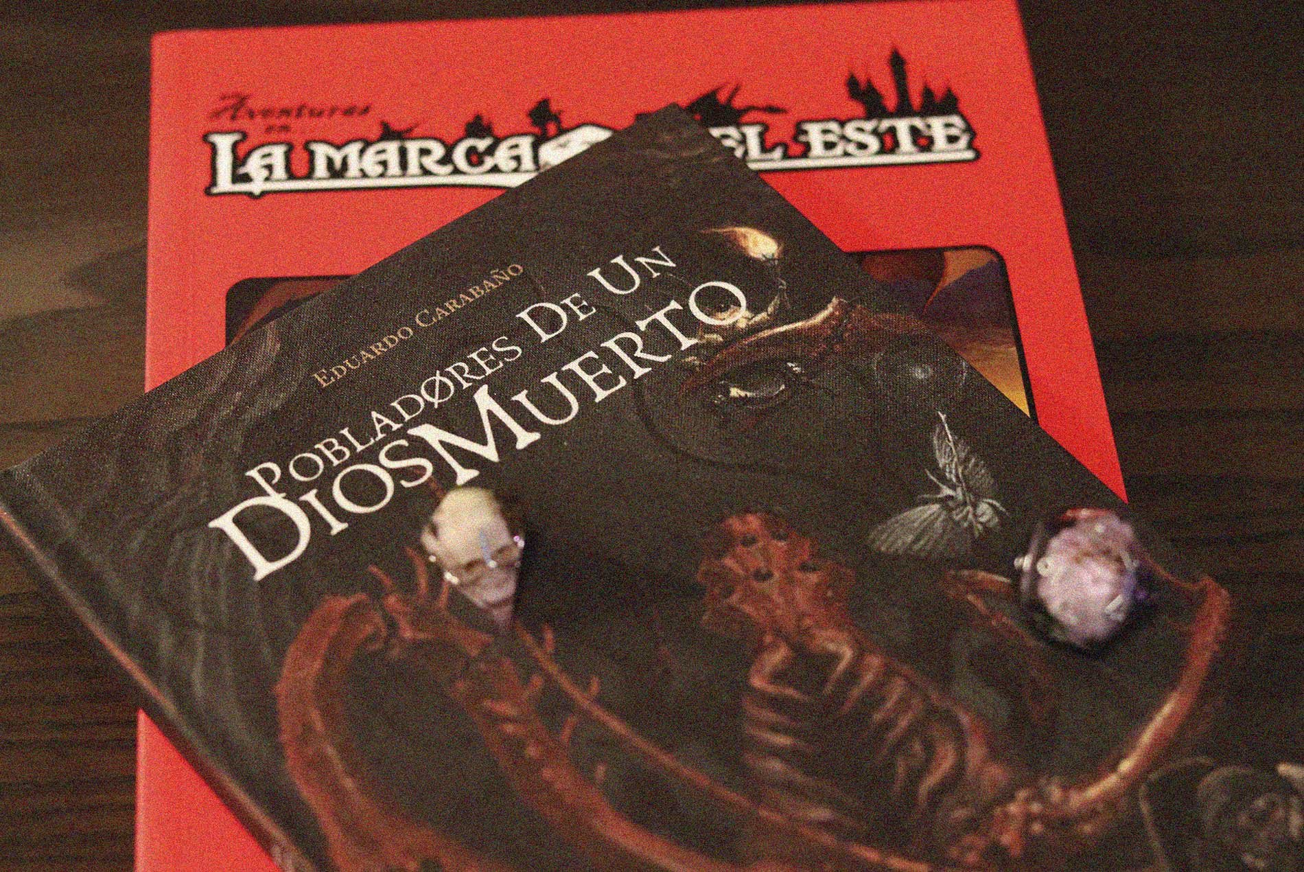 Fotografía del libro Pobladores de un Dios Muerto sobre la mesa, acompañado de dados y el manual de La Marca del Este al fondo.