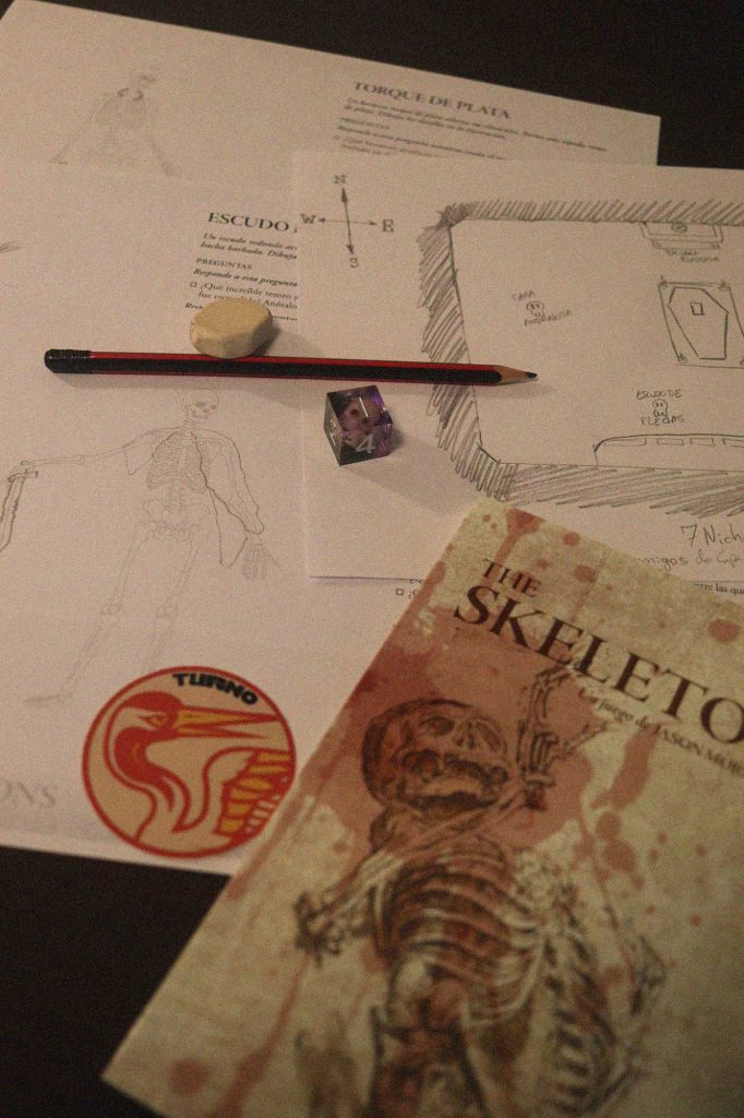 Mesa de juego de The Skeletons con libreto, hojas ilustradas de esqueletos, lápiz, goma y dado sobre papel de trabajo.