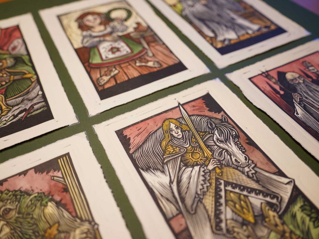 Varias ilustraciones tipo tarot en estilo linograbado dispuestas sobre una mesa verde, impresas en papel de algodón con bordes irregulares.