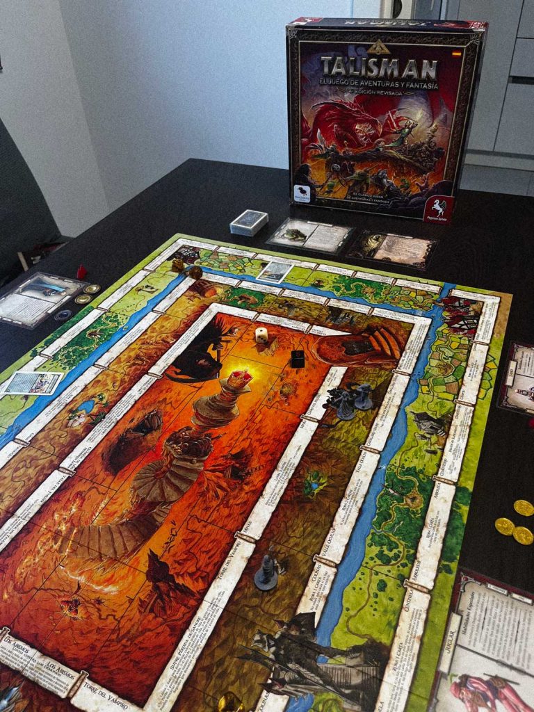 Partida del juego de mesa Talismán con el tablero desplegado, miniaturas, dados y la caja del juego al fondo