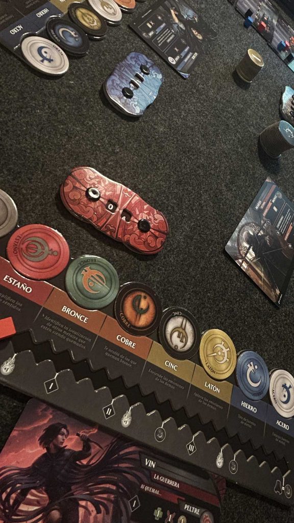 Tablero de metales y componentes de una partida de Nacidos de la Bruma junto a las cartas de Vin y Marsh.