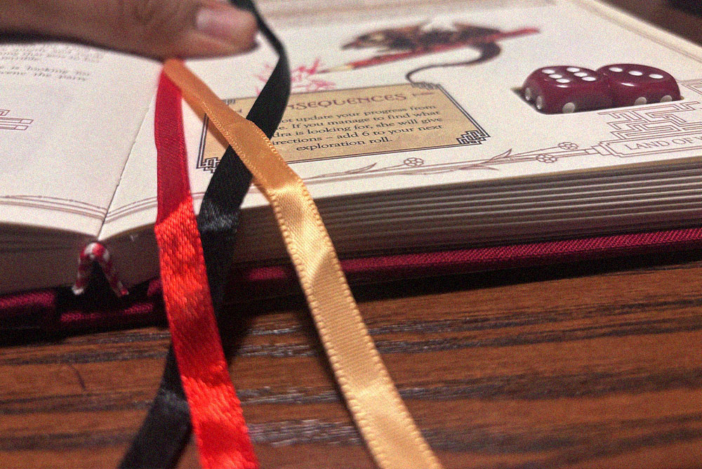 Libro de aventuras de rol abierto con un dado rojo y cintas de colores entre sus páginas.