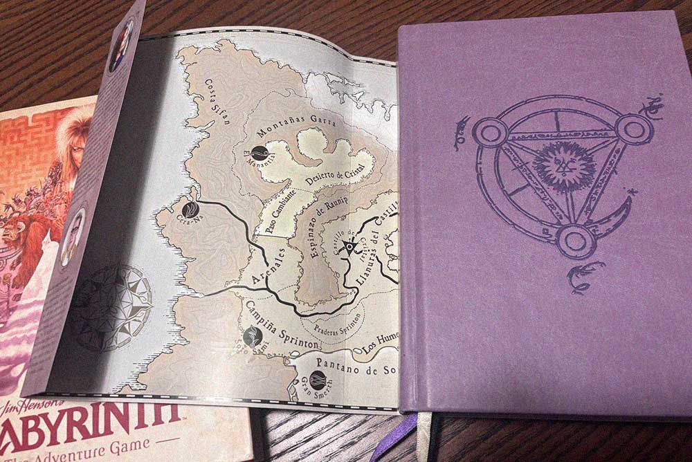 Mapa de fantasía abierto junto a un libro de aventuras de tapa morada con un símbolo esotérico, sobre una mesa de madera.